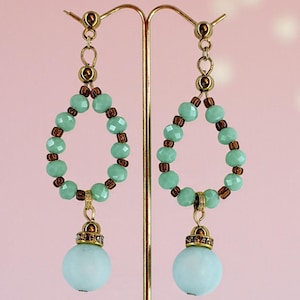 Peut inclure: Boucles d'oreilles pendantes élégantes. Elles présentent un motif en forme de goutte d'eau avec des perles vert clair et des entretoises marron. Une sphère bleu clair pend de chaque boucle d'oreille, ajoutant une touche de couleur et de sophistication.