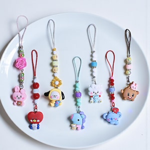 May include: A collection of colorful character keychains with beaded accents and attached loops. Each keychain features a different cartoon character in various colors, including pink, yellow, blue, and brown. The keychains are arranged on a white plate.