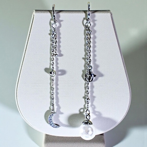 May include: A pair of silver dangle earrings. One earring features a crescent moon charm with small clear stones, and the other has a white pearl. Both earrings have a delicate chain design and are displayed on a white jewelry stand.