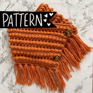 May include: Orange crochet neck warmer with a textured stitch and fringe. The neck warmer has two brown buttons.