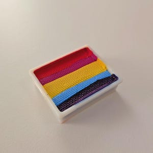 Può includere: Una tavolozza rettangolare di trucco per il viso con sei colori vivaci disposti a strisce orizzontali. I colori includono rosso, viola, giallo, blu e nero, contenuti in un contenitore di plastica bianca. Ideale per il trucco artistico.