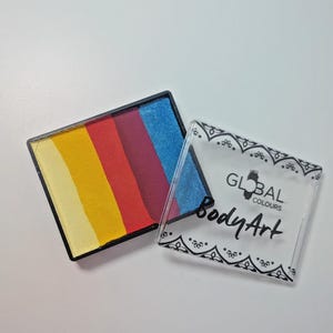 Può includere: Una tavolozza di colori per il trucco del viso con colori arcobaleno, con sezioni gialle, arancioni, rosse, viola e blu in un bordo nero. Accanto alla tavolozza c'è una copertura di plastica trasparente con la scritta "GLOBAL COLOURS Body Art".