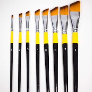 Puede incluir: Un juego de ocho pinceles angulados, dispuestos en una fila de menor a mayor tamaño. Cada pincel tiene un mango negro, una sección amarilla cerca de las cerdas y férulas de metal plateado. Las cerdas son de color marrón claro y los pinceles están numerados del 1 al 8.