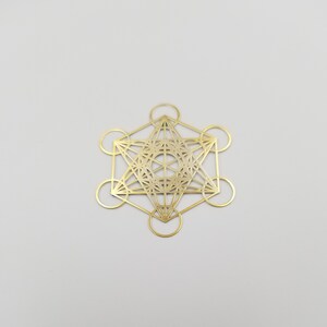 May include: A gold-colored metal pendant featuring a geometric design with a star shape in the center. The design is intricate and detailed, with multiple lines and circles.