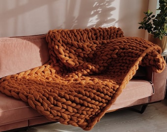 May include: A chunky knit throw blanket in a warm brown color draped over a pink sofa. The blanket is made of thick yarn and has a soft, cozy texture.