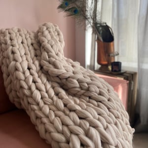 Puede incluir: Una manta de punto grueso de color beige neutro está sobre un sofá rosa. La gruesa textura tejida de la manta es el punto focal, lo que sugiere calidez y comodidad. Plumas de pavo real en un jarrón de cobre añaden un toque de elegancia.