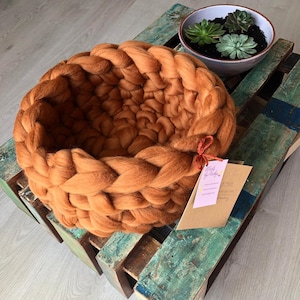 May include: A large, round, brown knitted basket made of chunky yarn. The basket is sitting on a wooden pallet with a small pot of succulents in the background. The basket is labeled "Work by Nicky".