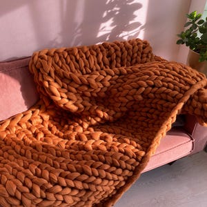 Puede incluir: Una manta de punto grueso en un cálido tono naranja, sobre un sofá rosa. La gruesa textura tejida de la manta es visible, creando una estética acogedora. La manta es de gran tamaño, perfecta para acurrucarse en ella.