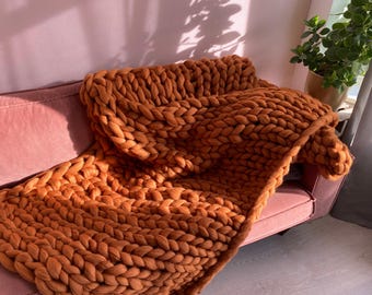 May include: A chunky knit blanket in a warm orange hue, draped over a pink sofa. The blanket's thick, woven texture is prominently displayed, creating a cozy and inviting aesthetic. The blanket is a large size, perfect for snuggling.