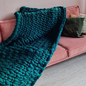 Puede incluir: Una manta de punto grueso de color verde azulado sobre un sofá rosa. El tejido grueso y texturizado de la manta es el punto focal, contrastando con el sofá liso. Cojines decorativos en tonos verdes y dorados añaden a la estética acogedora. La manta mide unos 152 cm de largo.