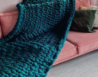 May include: A chunky knit teal blanket draped over a pink sofa. The blanket's thick, textured weave is the focal point, contrasting with the smooth sofa. Decorative pillows in shades of green and gold add to the cozy aesthetic. The blanket is approximately 60 inches long.