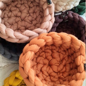 May include: Five woven pet beds in different colors, including teal, beige, pink, orange, and yellow. The beds are made of thick, chunky yarn and have a round shape.