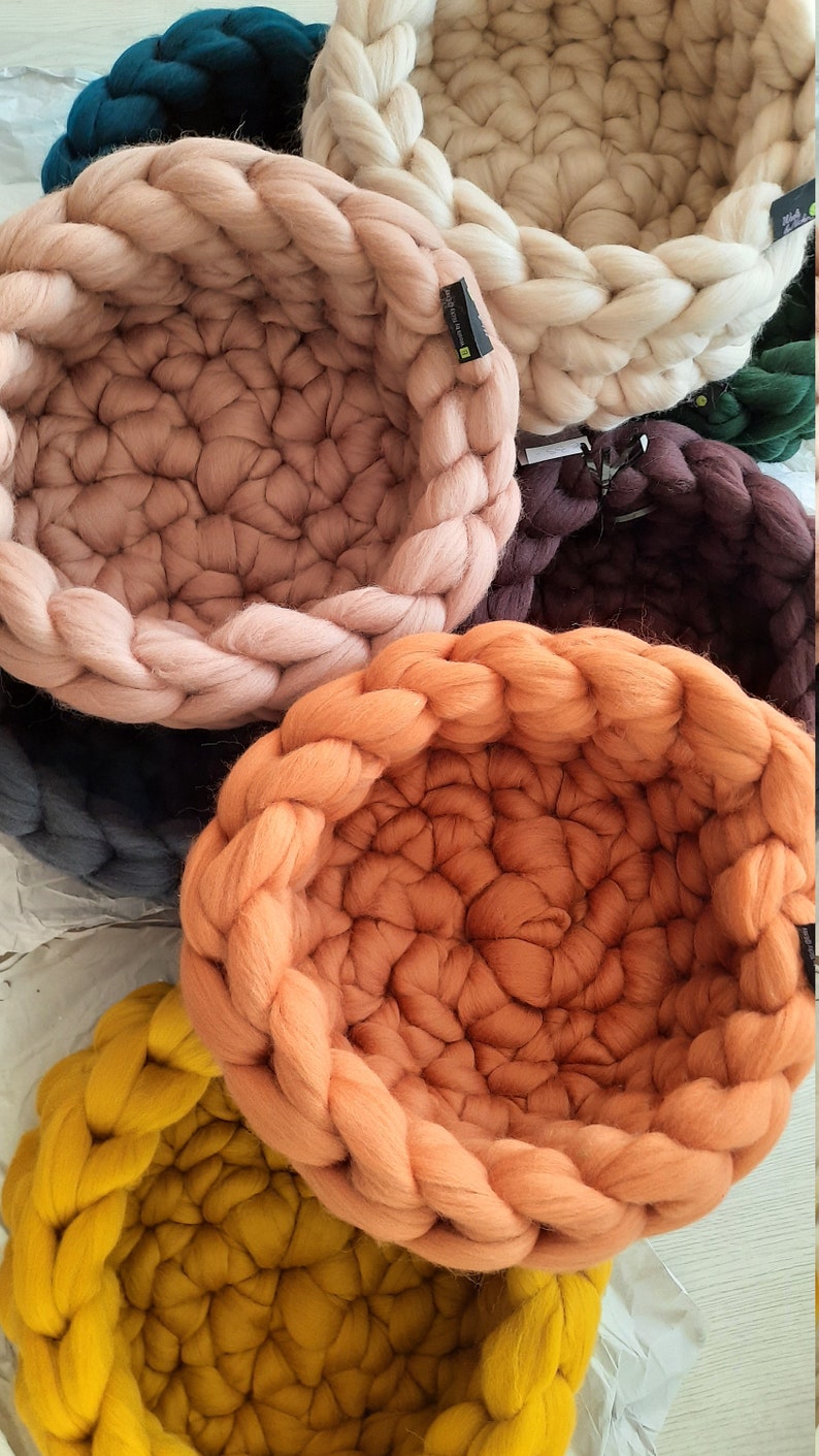 May include: Five woven pet beds in different colors, including teal, beige, pink, orange, and yellow. The beds are made of thick, chunky yarn and have a round shape.