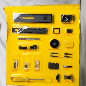 Puede incluir: Un paquete de cartón amarillo que contiene los componentes de una cámara de bolsillo u-build 110. El kit incluye un conjunto de lentes, botón disparador, rueda de avance de película, correa de muñeca, tornillos y un destornillador. El texto "u-build 110 POCKET CAMERA" es visible.