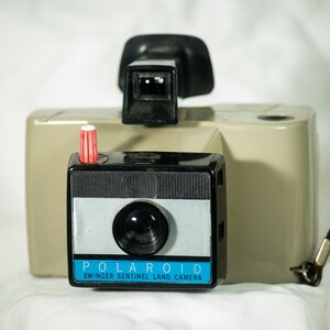 May include: A vintage Polaroid Swinger Sentinel Land Camera in beige with a black lens and a red button. The camera has a blue and white label that reads "POLAROID SWINGER SENTINEL LAND CAMERA".