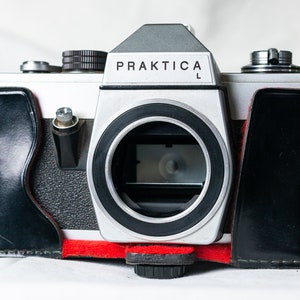 May include: A silver Praktica L camera with a black leather case. The camera has a lens cap and a strap.