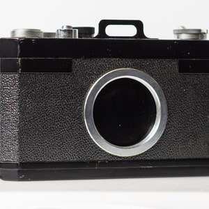 May include: A black vintage camera with a silver lens and a silver handle on top. The camera has a textured black leather covering.