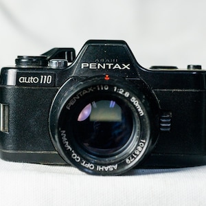Puede incluir: Una cámara de película negra Pentax auto 110 con una lente Pentax-110 1:2.8 50mm. La cámara tiene una etiqueta plateada auto 110 en la parte delantera.