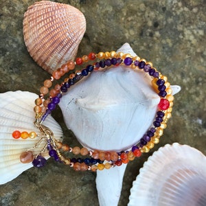 May include: A multi-strand bracelet with gold, orange, red, purple, and blue beads. The bracelet is displayed on a white seashell.