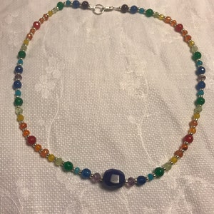 Könnte beinhalten: Eine Regenbogen-Perlenkette mit einem großen, dunkelblauen Edelstein-Anhänger. Die Kette besteht aus kleinen, runden Perlen in verschiedenen Farben, darunter Rot, Orange, Gelb, Grün, Blau und Lila.