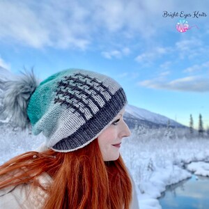 Op de afbeelding: Een gebreide muts met een gradiënt van groen, wit en grijs. De muts heeft een geometrisch patroon en een pluche pompon bovenop. De muts wordt gedragen door een persoon met rood haar, die in een besneeuwd landschap staat.