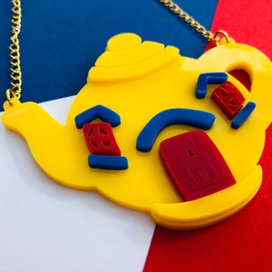 May include: A yellow teapot necklace with blue and red details. The teapot has a spout, handle, and a lid. The details include a window, a door, and a curved line.