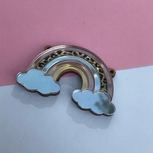 May include: An acrylic rainbow charm with a gold, pink and white colour scheme. The rainbow has a leopard print design and is accented with two mirrored clouds.