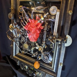 May include: Steampunk-style wall art featuring a red anatomical heart at the center, surrounded by gears, gauges, and tubes. The artwork is framed in a dark, metallic finish, with a green-lit element at the top. A unique, industrial-inspired piece.