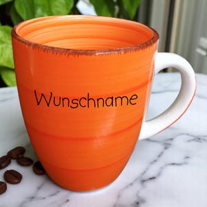 Könnte beinhalten: Orangefarbene Keramik-Tasse mit weißem Henkel und dem Wort "Wunschname" in Schwarz. Die Tasse hat horizontale Streifen und einen braunen Rand. Kaffeebohnen sind in der Nähe der Tasse verstreut.