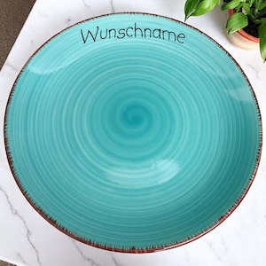 Könnte beinhalten: Eine große, türkisfarbene Keramikplatte mit Spiralmuster. Die Platte hat einen braunen Rand und das Wort "Wunschname" in schwarzer Schrift. Die Platte steht auf einer weißen Marmoroberfläche.