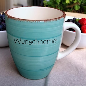 Könnte beinhalten: Eine türkisfarbene Keramiktasse mit horizontalen Streifen und dem Wort "Wunschname" in schwarzer Schrift. Die Tasse hat einen weißen Innenraum und Henkel, mit einem braunen Rand. Beeren sind im Hintergrund zu sehen.
