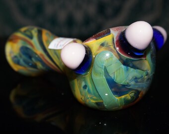 May include: A colorful glass pipe with a swirl design and three white and blue eyes. The pipe is made of glass and has a green, blue, and yellow color scheme.