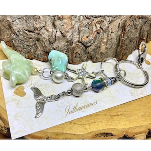 May include: A collection of keychains with a mermaid theme. The keychains feature a variety of charms, including a green dolphin, a starfish, and a mermaid tail. The keychains are attached to silver key rings. The text "Intheweaves" is visible.
