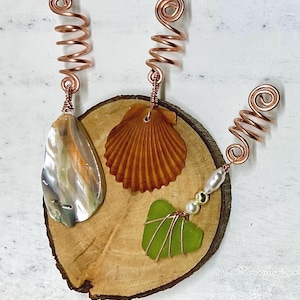 May include: Handmade copper wire hair accessories featuring a pearlescent shell, a brown seashell, and a green sea glass piece. The accessories are attached to a wooden slice, showcasing a natural, beach-inspired theme.