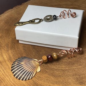 May include: Two handmade hair accessories. One features a seashell, wooden beads, and copper wire. The other has a metal charm, black bead, and copper wire. Both are on a white box, set on a wooden surface.