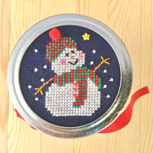 Pode incluir: Um desenho de boneco de neve em ponto cruz sobre um fundo azul escuro, inserido numa tampa de metal prateado. O boneco de neve usa um chapéu e cachecol vermelhos e verdes, com uma estrela amarela e flocos de neve brancos. Uma fita vermelha é visível.