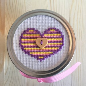 Peut inclure: Un cœur brodé en point de croix sur un fond de tissu blanc, avec des rayures violettes et jaunes. Le cœur a un bouton en bois au centre.