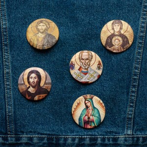 Könnte beinhalten: Fünf runde Anstecknadeln mit religiösen Ikonen, die Jesus, Maria und andere Figuren darstellen. Die Anstecknadeln werden auf einem blauen Jeanshintergrund gezeigt. Die Anstecknadeln weisen eine Vielzahl von Farben auf, darunter Gold, Braun, Blau und Rot.