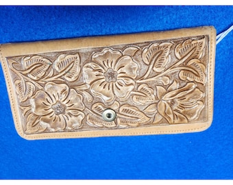 Puede incluir: Cartera de cuero color canela con un diseño floral en relieve. La cartera tiene un cierre de botón a presión y es de forma rectangular. Las flores y hojas en relieve son detalladas y crean una superficie texturizada. El fondo es azul liso.