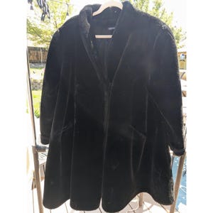 May include: A black faux fur coat featuring a stand-up collar, long sleeves, and a flared silhouette. The coat has a smooth, plush texture throughout, a single front closure, and a simple, unadorned design that falls to the mid-thigh area.