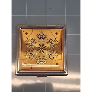 May include: A vintage silver compact with a gold-toned floral design on the lid. The compact has a hinged lid and a mirror inside.