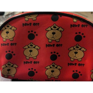 Pode incluir: Bolsa de cosméticos de tecido vermelho com um padrão repetido de cães de desenho animado marrons com o texto "paws off" e pegadas pretas.