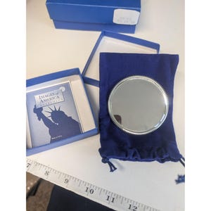 Puede incluir: Un espejo compacto redondo de color plateado con un borde con cuentas, apoyado en una bolsa de terciopelo azul oscuro. El espejo está junto a una caja azul que contiene un folleto con la Estatua de la Libertad en la portada. La caja está abierta.