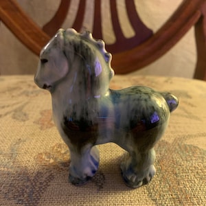 Puede incluir: Una figurita de caballo de cerámica con un esmalte azul y verde. El caballo tiene una larga melena y cola y está de pie sobre sus cuatro patas.