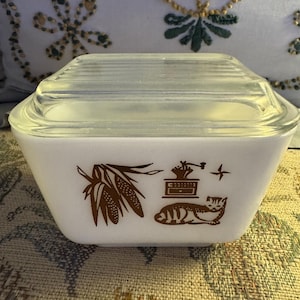 May include: A white, square-shaped Pyrex dish with a clear glass lid. The dish features brown graphic designs of corn, a coffee grinder, a star, and a cat. The dish is sitting on a patterned surface.