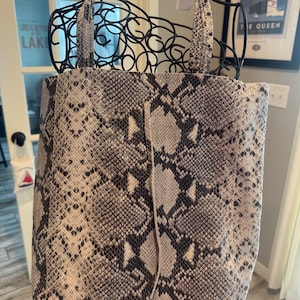 May include: A tote bag featuring a grey, black, and white snakeskin pattern. The bag has two matching top handles and a thin, vertical leather cord detail centred on the front panel. The material has a textured finish throughout the exterior.