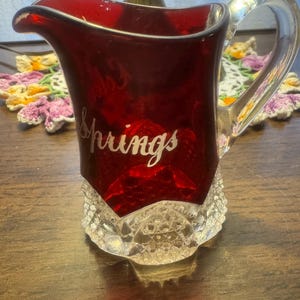 Puede incluir: Una jarra de cristal roja vintage con asa y base de cristal transparente. La palabra "Springs" está escrita en cursiva en la parte delantera. La base tiene un patrón de diamantes.
