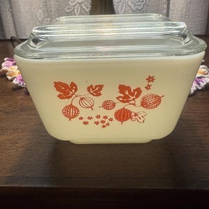 May include: A vintage, off-white Pyrex bowl with a clear glass lid. The bowl features a red gooseberry design. The bowl is a classic kitchenware item, perfect for serving or storing food. The bowl is in good condition.