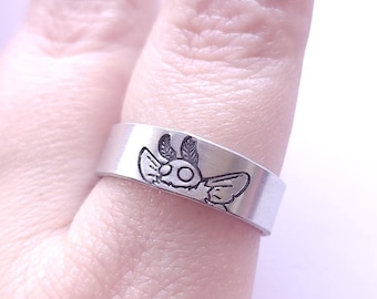 Puede incluir: Un anillo de plata con un pequeño diseño grabado de una criatura con alas. El anillo se lleva en un dedo.