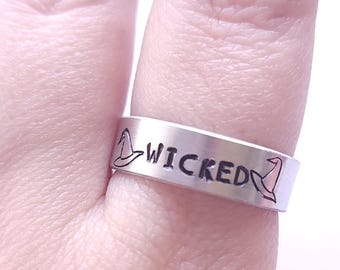 May include: A silver ring with the word "WICKED" engraved on it. The ring is decorated with two witch hat symbols on either side of the word.